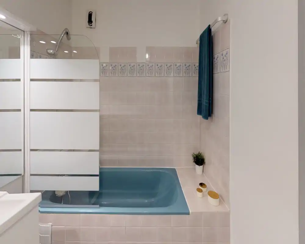 Clean, well-lit bathroom featuring a blue bathtub with a partial frosted glass shower screen, tiled walls with decorative border, and a towel on the rail.