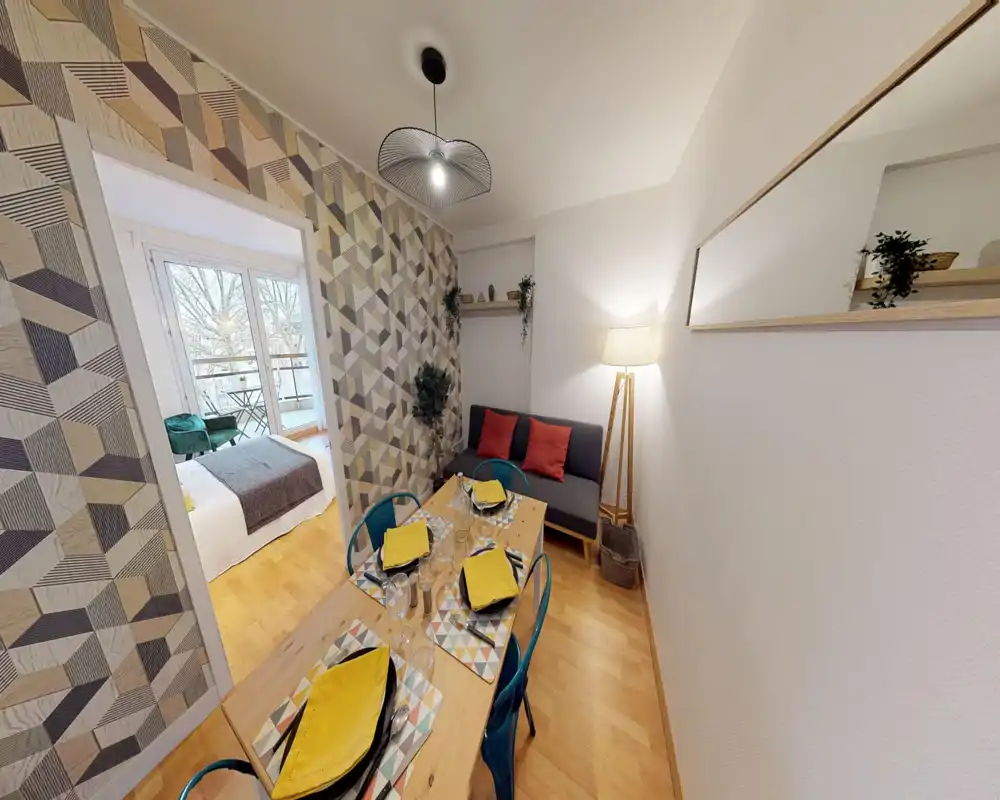 Bright, styled living/dining area with a wooden table set for four, patterned accent wall and a small sofa — an inviting space that opens to a bedroom with balcony.