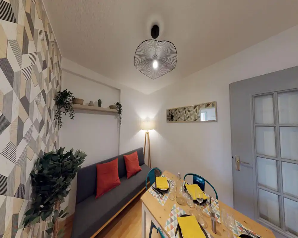 A cozy, well-styled living/dining corner with a dark sofa, colorful cushions, patterned wallpaper and a small wooden dining table set for four. Warm lighting and decorative plants create a welcoming atmosphere ideal for short-term rentals.