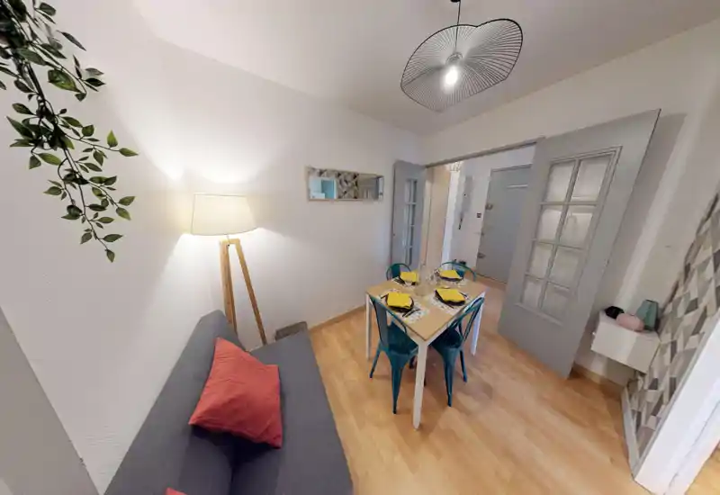 Bright, well-styled small living/dining area with a sofa, side lamp and a four-seat dining table set for guests. The neutral walls and warm wooden floor give a cozy, modern feeling ideal for short-term rentals.