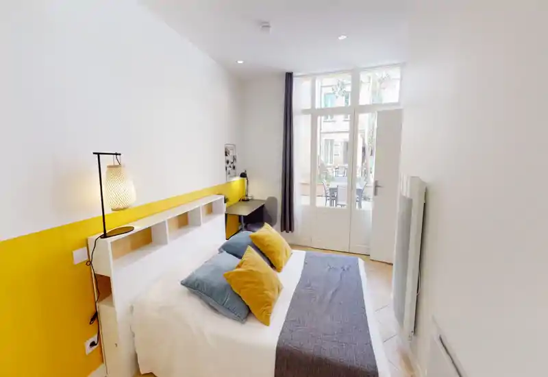 Chambre lumineuse et soignée avec lit double, coussins colorés et grandes fenêtres donnant sur un espace extérieur. Décor moderne et minimal avec un coin bureau et éclairage chaleureux.