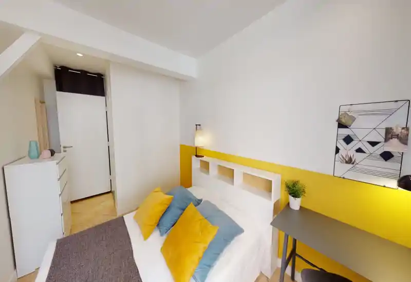 Chambre moderne et lumineuse avec lit double bien fait, coussins jaunes et bleus, lampe de chevet et petit bureau — idéale pour les locations courte durée stylées.