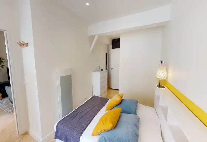 Chambre moderne et lumineuse avec un lit double bien fait, coussins colorés et une lampe de chevet chaleureuse. Murs blancs, commode et éléments soignés offrent une ambiance cosy, idéale pour une annonce locative.