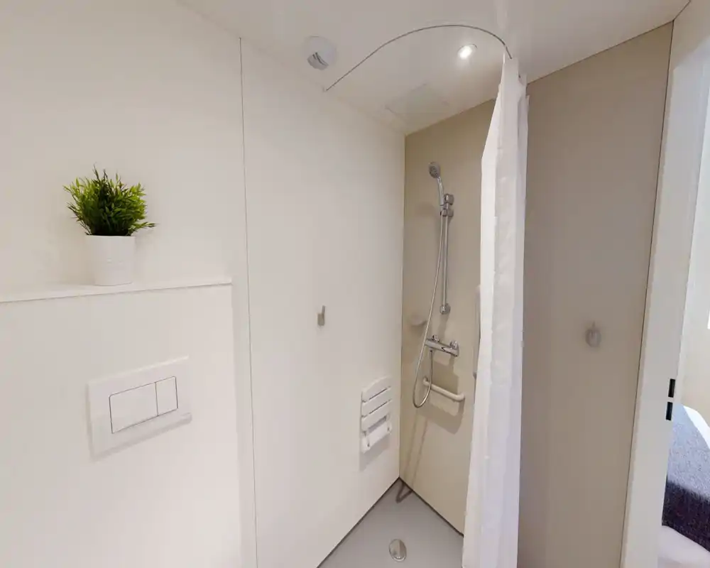 Zona de ducha compacta y accesible con ducha de mano, asiento abatible y cortina blanca; baño luminoso y minimalista, pensado para un uso cómodo.