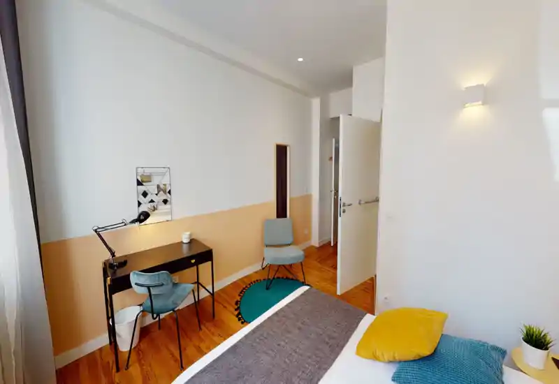 Dormitorio luminoso y moderno con cama cómoda en primer plano, mantas y cojines texturizados, un pequeño escritorio junto a la ventana y una alfombra acogedora — ideal para una persona o pareja.