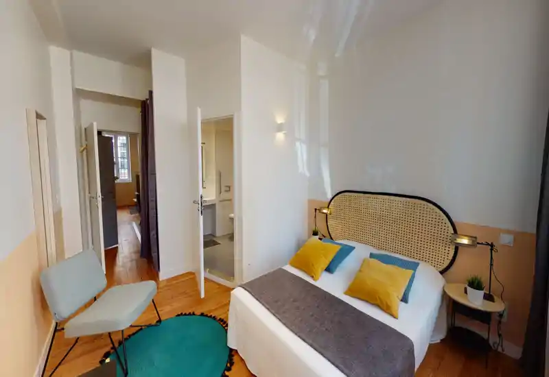 Dormitorio luminoso y moderno con cabecero de mimbre, ropa de cama blanca, cojines coloridos y mesita con lámpara y planta. Suelo de madera, butaca y acceso directo al baño en suite.