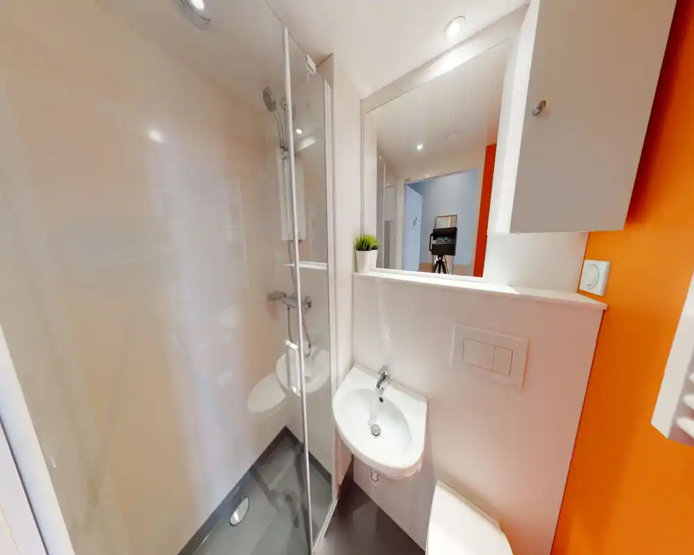 Salle de bain moderne et compacte avec cabine de douche d'angle, lavabo mural et toilettes; un grand miroir et un bon éclairage donnent une impression propre et fonctionnelle.