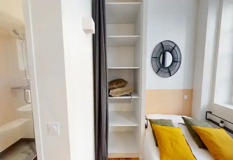 Coin chambre lumineux et bien mis en scène avec un lit double garni de coussins verts et moutarde, un miroir mural rond décoratif et des étagères ouvertes pour le rangement. La lumière naturelle renforce l'atmosphère chaleureuse et accueillante.