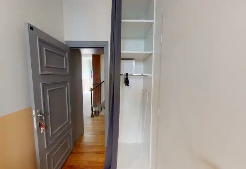 Petit espace d'entrée avec une porte grise décorative ouverte, parquet en bois et placard ouvert avec quelques cintres ; murs neutres et lumière naturelle depuis la pièce adjacente.