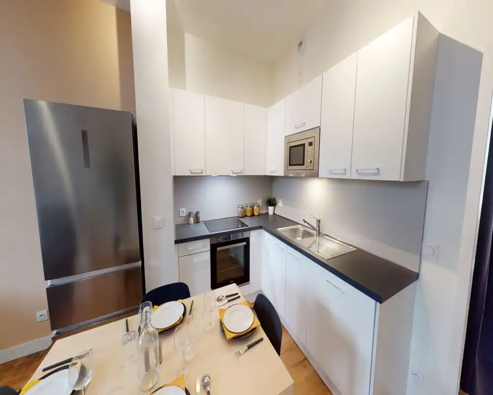 Cuisine moderne et lumineuse avec armoires blanches, appareils intégrés, évier en inox et réfrigérateur; une petite table dressée pour quatre personnes crée un espace convivial et fonctionnel.