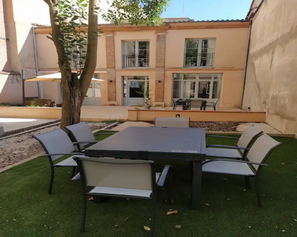 Cour intérieure ensoleillée avec un ensemble de repas extérieur moderne sur gazon artificiel, un arbre fournissant de l'ombre et la façade d'un bâtiment de deux étages en arrière-plan — idéal pour les repas en plein air.