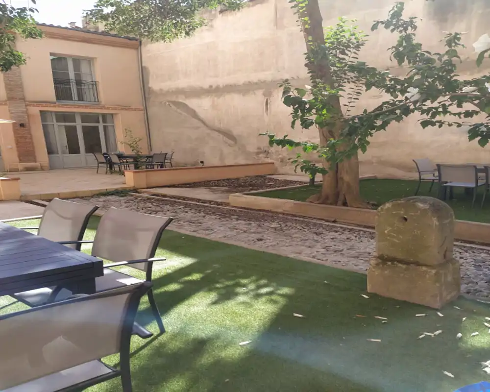 Cour privée ensoleillée avec coin repas extérieur, gazon synthétique et arbre mature — idéale pour se détendre ou dîner en plein air.
