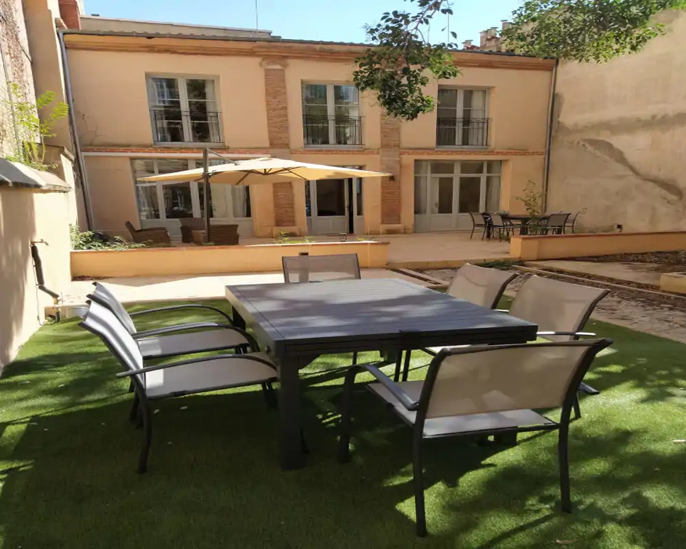 Cour intérieure ensoleillée avec table et chaises sur gazon synthétique, patio avec parasol et sièges supplémentaires — idéal pour repas en plein air et détente.