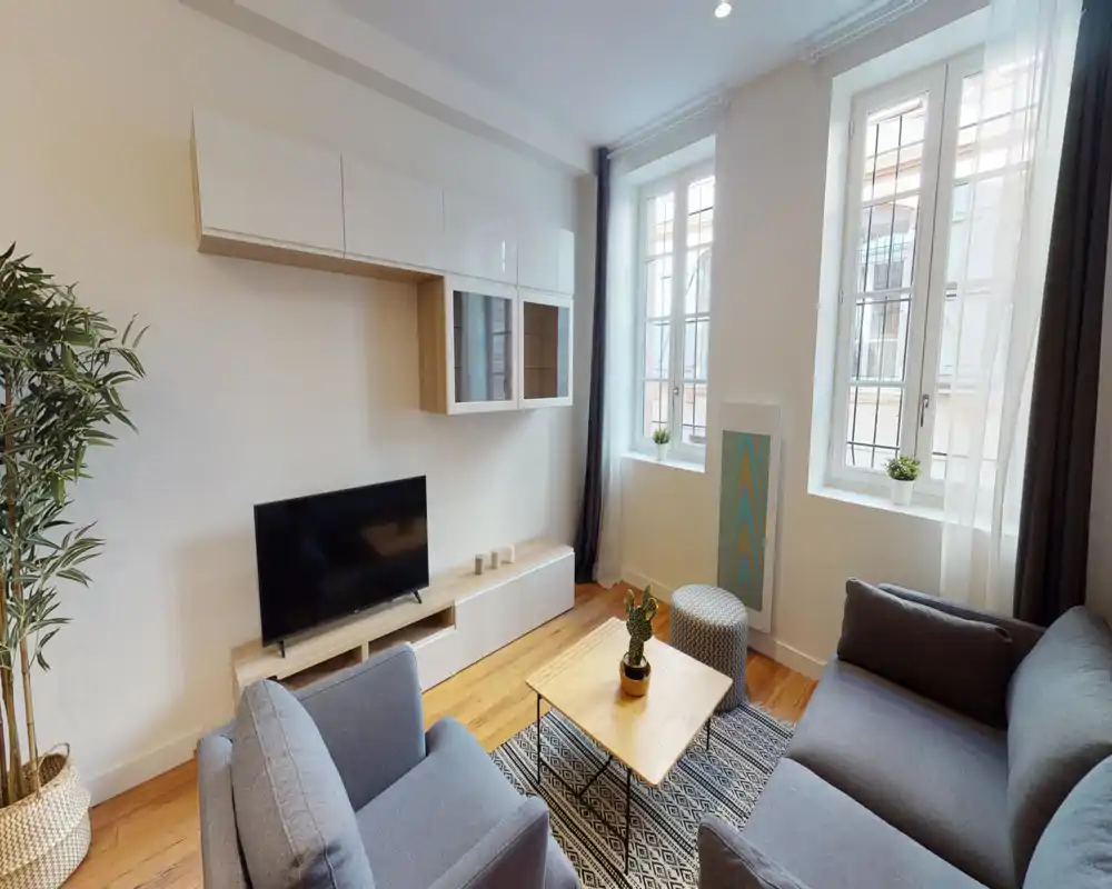 Salon lumineux et soigné avec deux canapés gris confortables, un tapis à motifs et une table basse; de grandes fenêtres éclairent la pièce et des meubles muraux conservent un aspect ordonné.