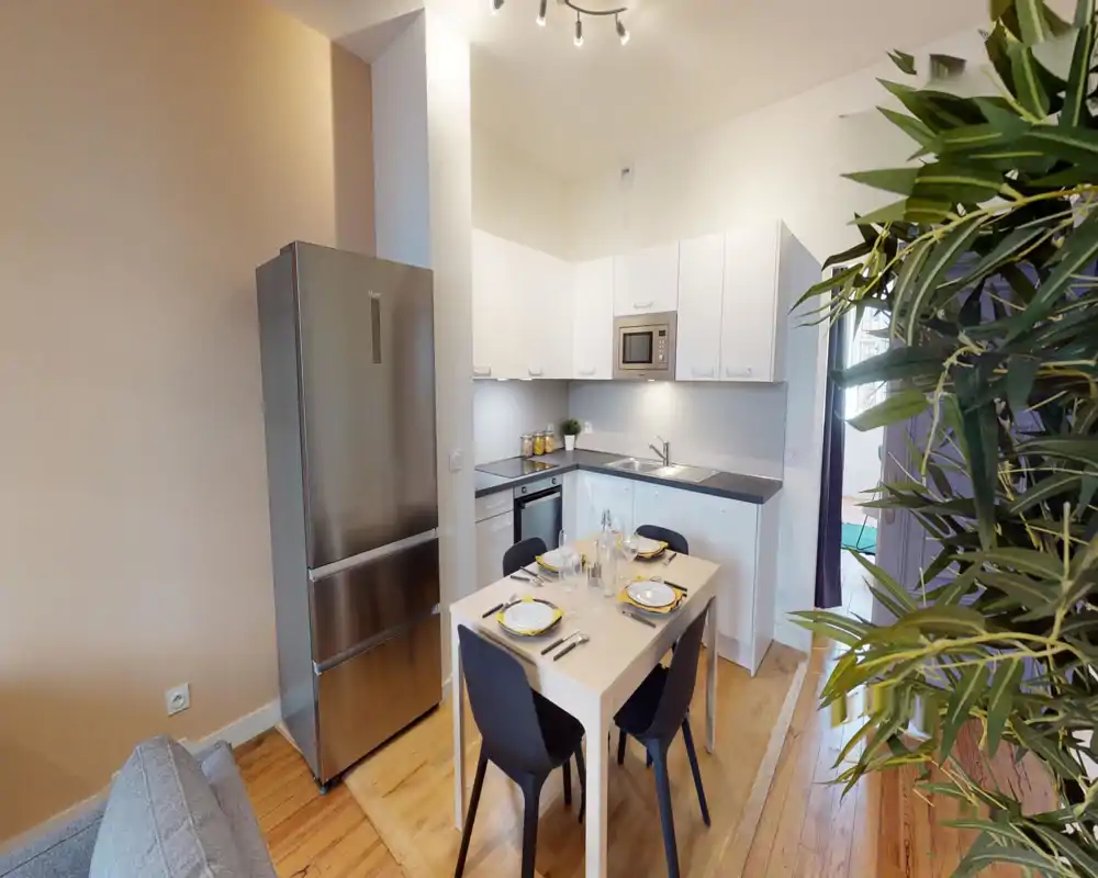 Cuisine ouverte moderne avec meubles blancs, appareils en acier inoxydable et petite table à manger pour quatre. Espace lumineux et bien mis en scène avec parquet et plante pour une touche chaleureuse.