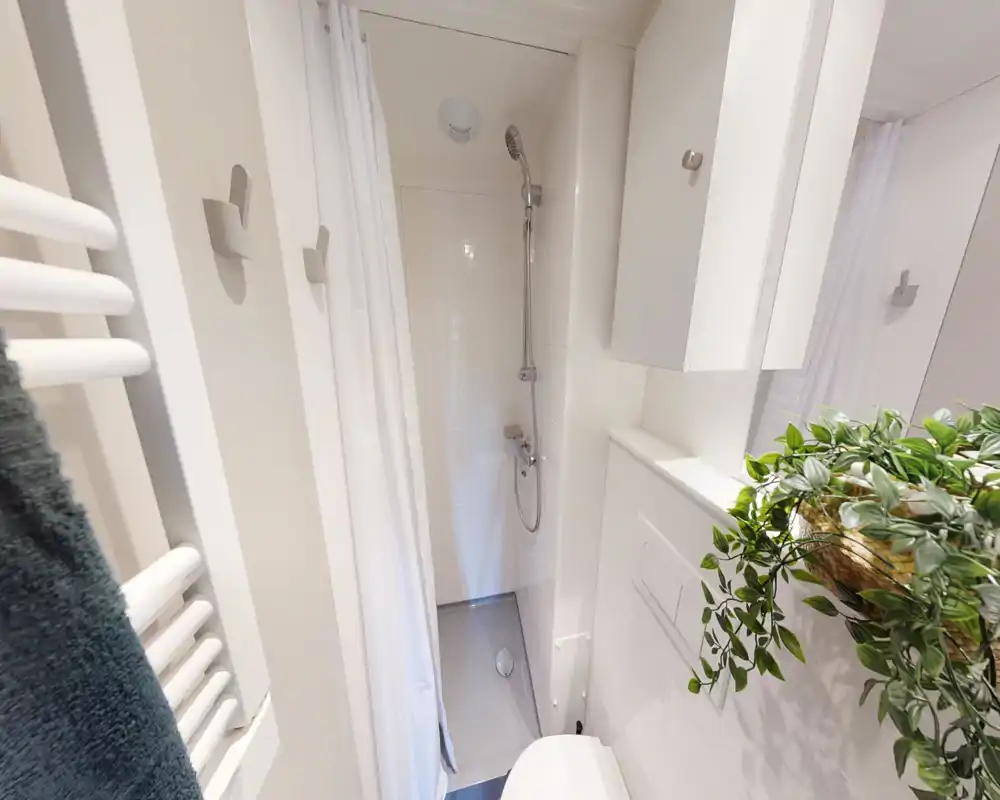Bagno compatto e moderno con doccia walk-in, piastrelle bianche, pensile a parete e una pianta decorativa per un tocco fresco.