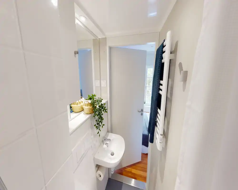 Bagno compatto e ben illuminato con piastrelle bianche, piccolo lavabo a muro, scalda-asciugamani e tenda per la doccia. Finiture moderne e cesti intrecciati per un tocco decorativo.