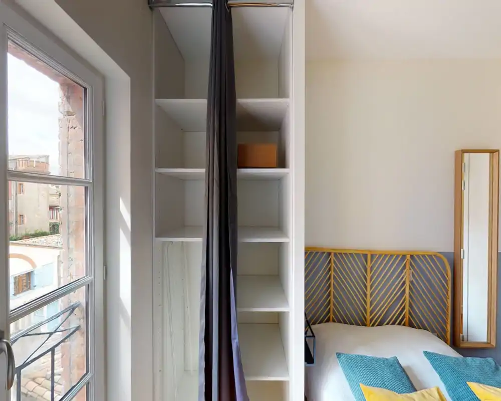 Camera da letto luminosa e minimale con testiera in motivo rattan, biancheria bianca e cuscini colorati; una scaffalatura integrata e una finestra a tutta altezza garantiscono luce e spazio di archiviazione.