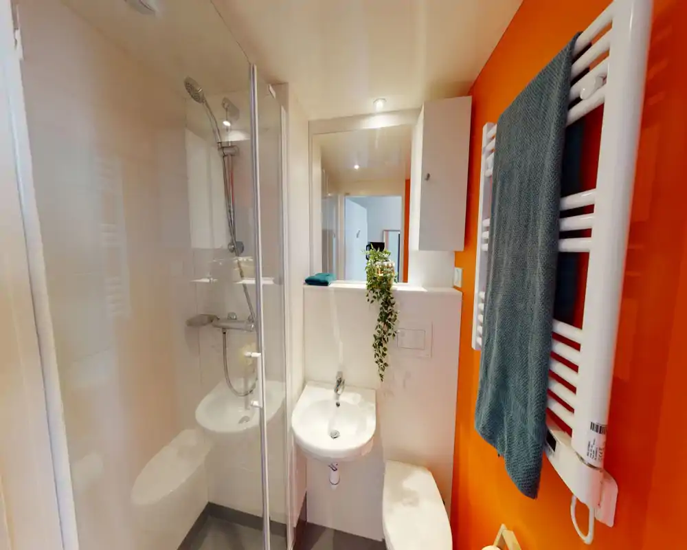 Bagno compatto e moderno con cabina doccia in vetro, lavabo a muro, WC e scaldasalviette su una parete arancione vivace. Linee pulite e una pianta appesa donano un aspetto fresco e curato.