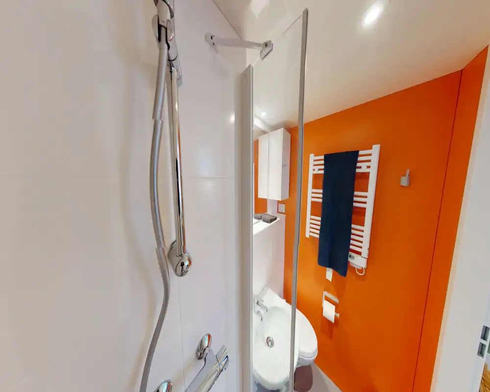 Baño moderno y compacto con ducha y lavabo visibles, pared naranja llamativa y toallero calefactado. Espacio limpio y bien iluminado, ideal para estancias cortas.