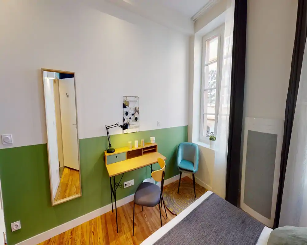 Rincón de dormitorio luminoso y bien decorado con borde de la cama visible, escritorio de madera compacto, lámpara de lectura y butaca junto a la ventana — paredes verdes y blancas y suelo de madera que aportan calidez.