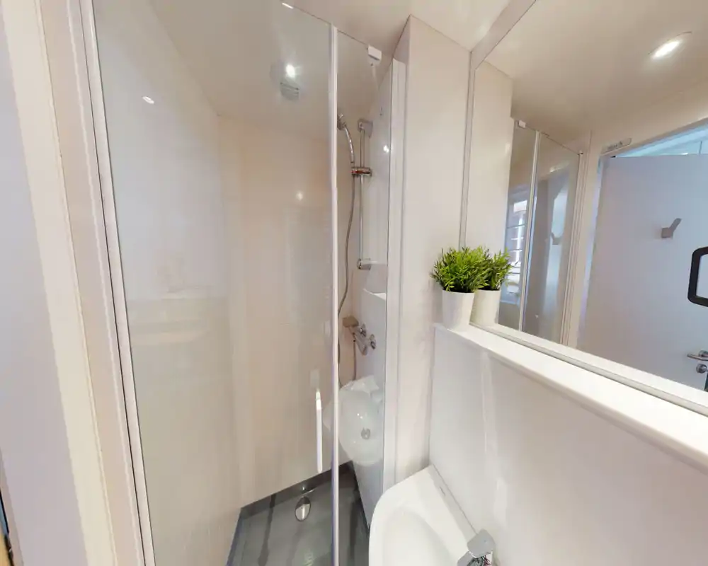 Baño moderno y compacto con ducha acristalada, grifería mural y un lavabo pequeño; acabados blancos limpios y una planta decorativa junto al espejo.