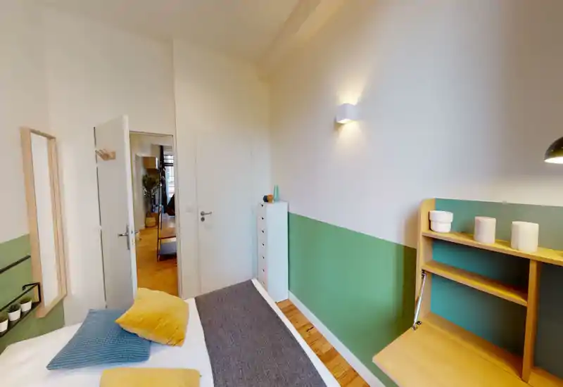 Camera da letto luminosa e ben curata con letto matrimoniale visibile, cuscini decorativi e una scrivania compatta in legno; parete bicolore verde e parquet caldo creano un’atmosfera accogliente.