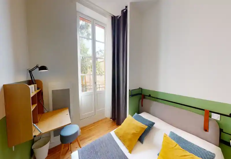 Camera da letto luminosa e ben arredata con un comodo letto matrimoniale, cuscini decorativi e pavimento in legno. Una grande finestra garantisce luce naturale e una piccola scrivania pieghevole offre spazio di lavoro pratico.