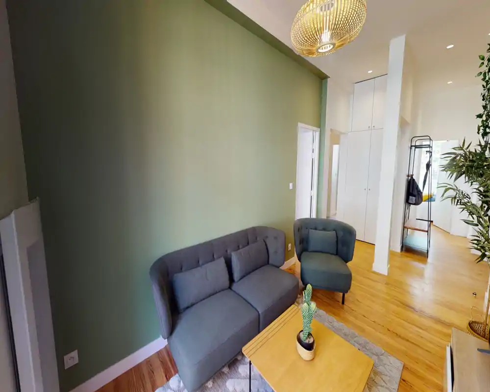 Soggiorno moderno e accogliente con divano grigio compatto e poltrona abbinata su una parete verde tenue, pavimenti in legno caldo e luce naturale — perfetto per rilassarsi o ricevere ospiti.
