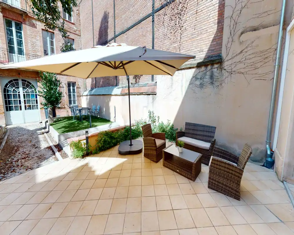 Cortile privato soleggiato con pavimento piastrellato, set di sedute in rattan e grande ombrellone — ideale per relax all'aperto e piccoli incontri.