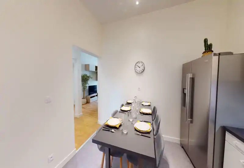 Zona pranzo elegante con tavolo apparecchiato per sei persone, tavolo e sedie grigie moderne e un grande frigorifero in acciaio visibile di lato — luminosa, minimal e pronta per gli ospiti.