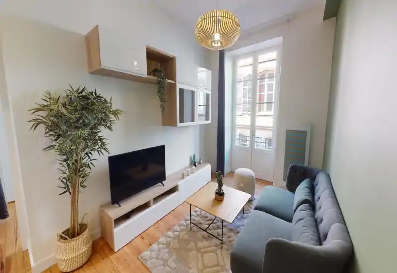 Soggiorno luminoso e moderno con divano capitonné comodo, tavolino basso in legno, mobile TV e ampia finestra che illumina l'ambiente. Pavimenti in legno, piante e tessili morbidi creano un'atmosfera accogliente.