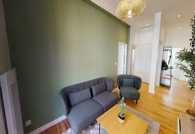 Soggiorno moderno e accogliente con divano grigio compatto e poltrona abbinata su una parete verde tenue, pavimenti in legno caldo e luce naturale — perfetto per rilassarsi o ricevere ospiti.