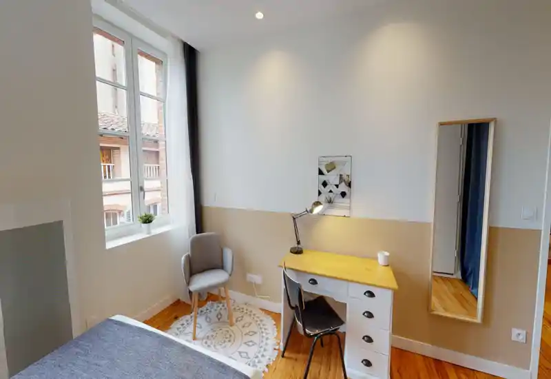 Bright, cozy bedroom corner with a visible bed edge, a desk and a reading chair near a large window offering natural light.