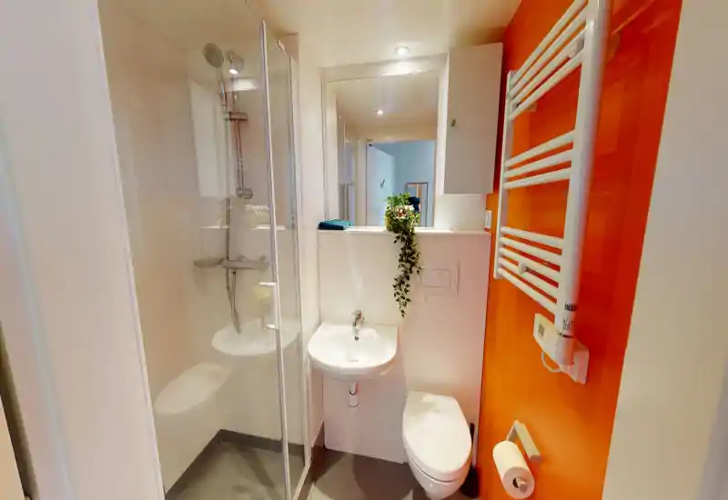 Compact modern bathroom with a walk-in shower, wall-mounted toilet and small sink; bright lighting and a warm orange accent wall add character.