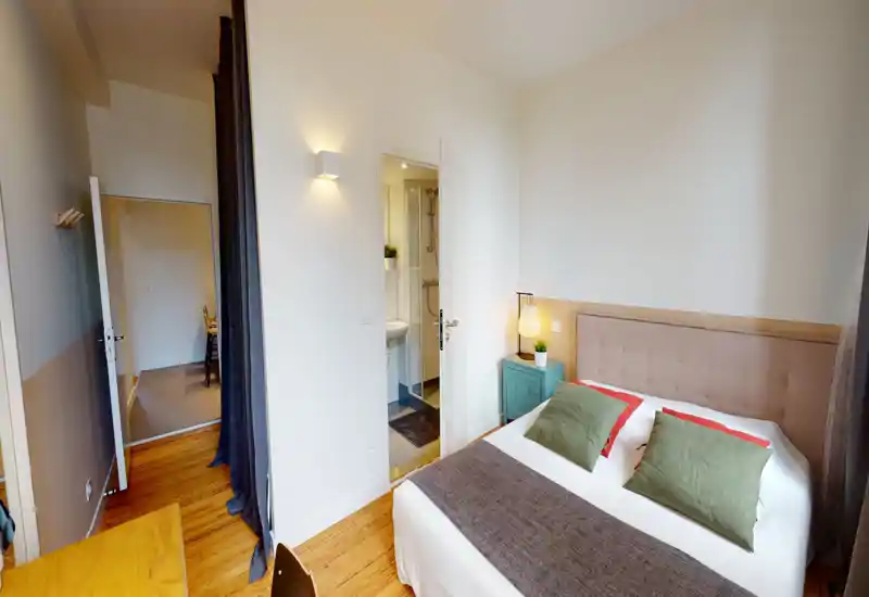 Chambre lumineuse et bien aménagée avec un grand lit, table de chevet et lampe. Décor neutre et sol en bois pour une ambiance chaleureuse et accueillante, idéale pour la location.