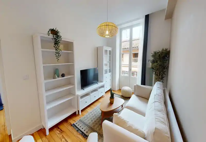 Salon lumineux et élégant avec un canapé blanc confortable, meuble TV, table basse et tapis décoratif. Une grande fenêtre apporte une belle lumière naturelle et le parquet chaud donne du caractère.