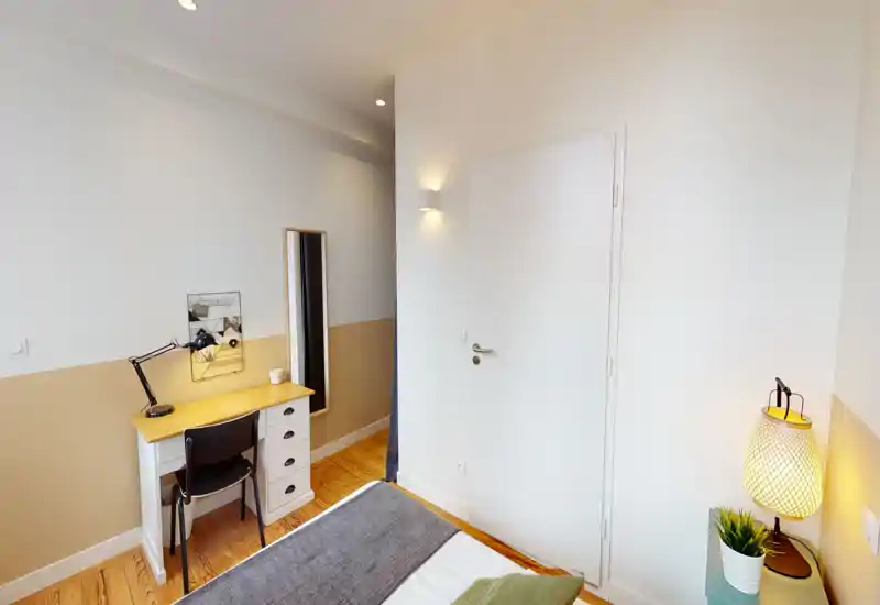 Chambre chaleureuse et bien éclairée avec le bord du lit visible, sol en bois et un petit bureau. Décor moderne et épuré, éclairage chaud pour une ambiance accueillante.