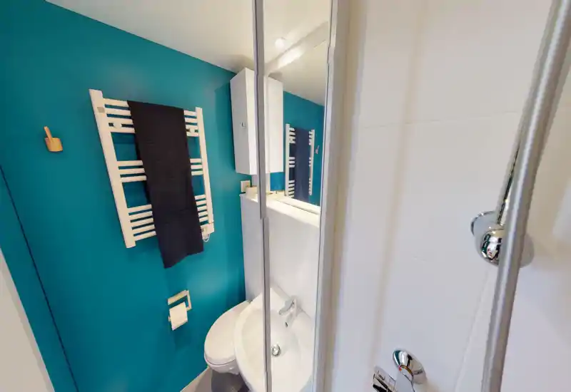 Bagno moderno compatto con lavabo bianco, WC e rubinetteria cromata, parete turchese vivace e scalda-asciugamani.