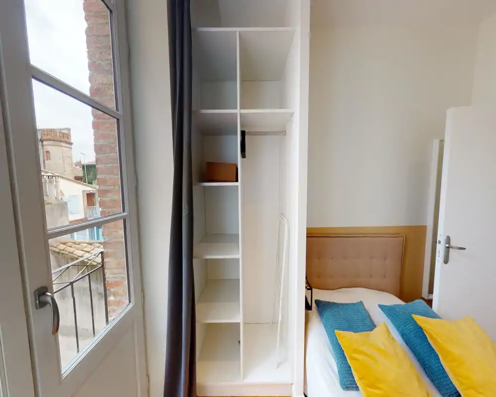 Chambre lumineuse et bien mise en scène avec une tête de lit capitonnée, coussins colorés et une penderie intégrée à côté d'une fenêtre à la française apportant la lumière naturelle.