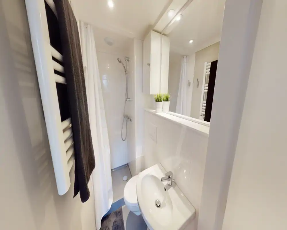 Salle de bains compacte et moderne avec lavabo mural, WC, douche avec rideau, sèche-serviettes et armoire miroir — lumineuse, propre et bien éclairée.