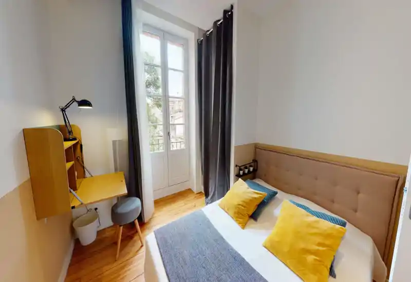 Chambre chaleureuse et lumineuse avec tête de lit capitonnée, coussins colorés et lit placé près d'une haute fenêtre. Un bureau mural compact et un tabouret complètent cet espace pratique et soigné.