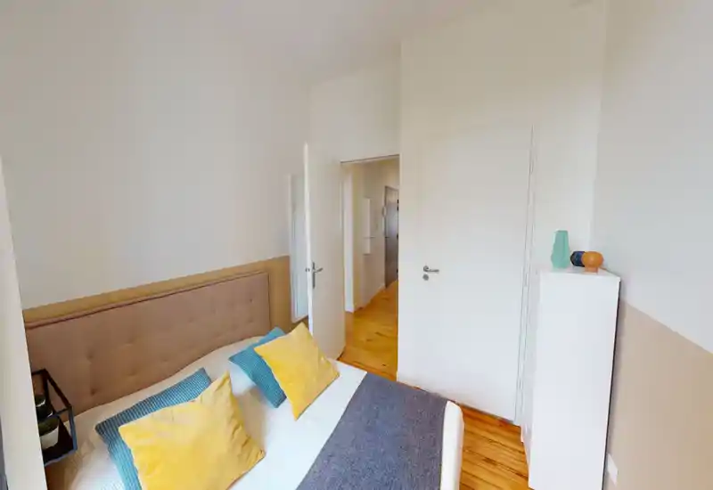 Chambre lumineuse et accueillante avec tête de lit capitonnée, coussins colorés et parquet en bois ; bien éclairée et décorée avec goût.