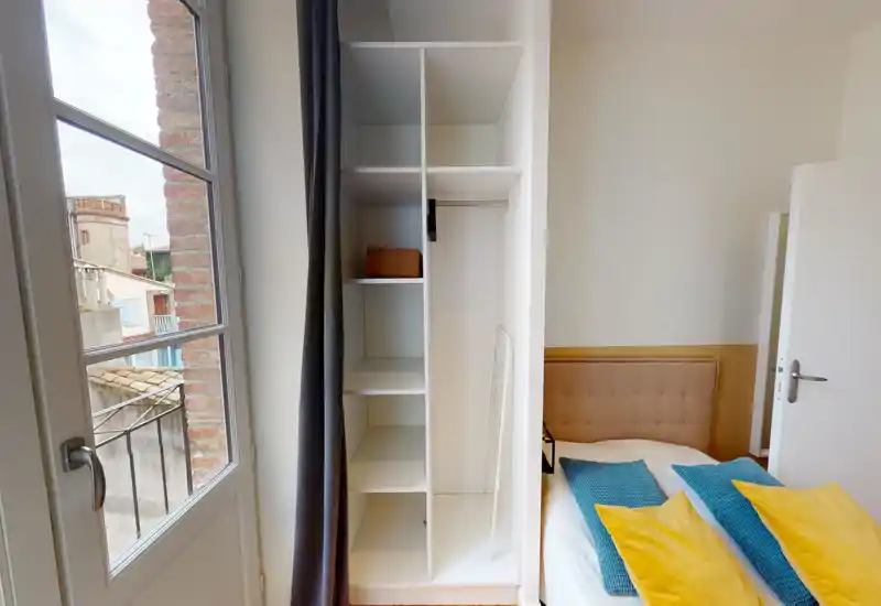 Chambre lumineuse et bien mise en scène avec une tête de lit capitonnée, coussins colorés et une penderie intégrée à côté d'une fenêtre à la française apportant la lumière naturelle.