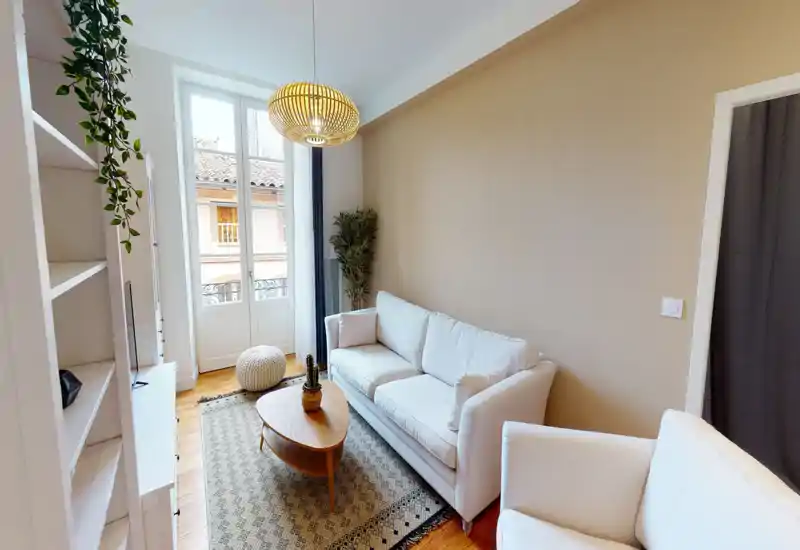 Soggiorno luminoso e accogliente con divano chiaro, tappeto decorato, tavolino in legno e ampia finestra che lascia entrare luce naturale — perfetto per rilassarsi o ricevere ospiti.