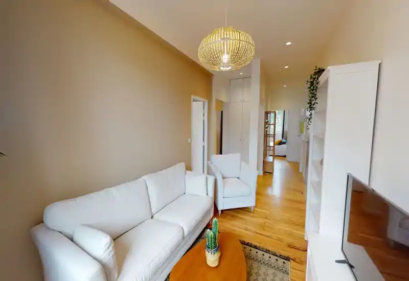 Soggiorno luminoso e ben arretrato con divano e poltrona bianchi, pavimenti in legno e un lampadario decorativo — accogliente e moderno.