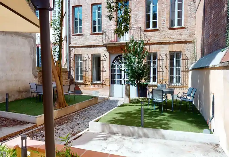 Cortile soleggiato con area esterna arredata: piccolo terrazzo con tavoli e sedie, prato sintetico e alberi davanti a una facciata in mattoni — perfetto per godersi l'esterno.