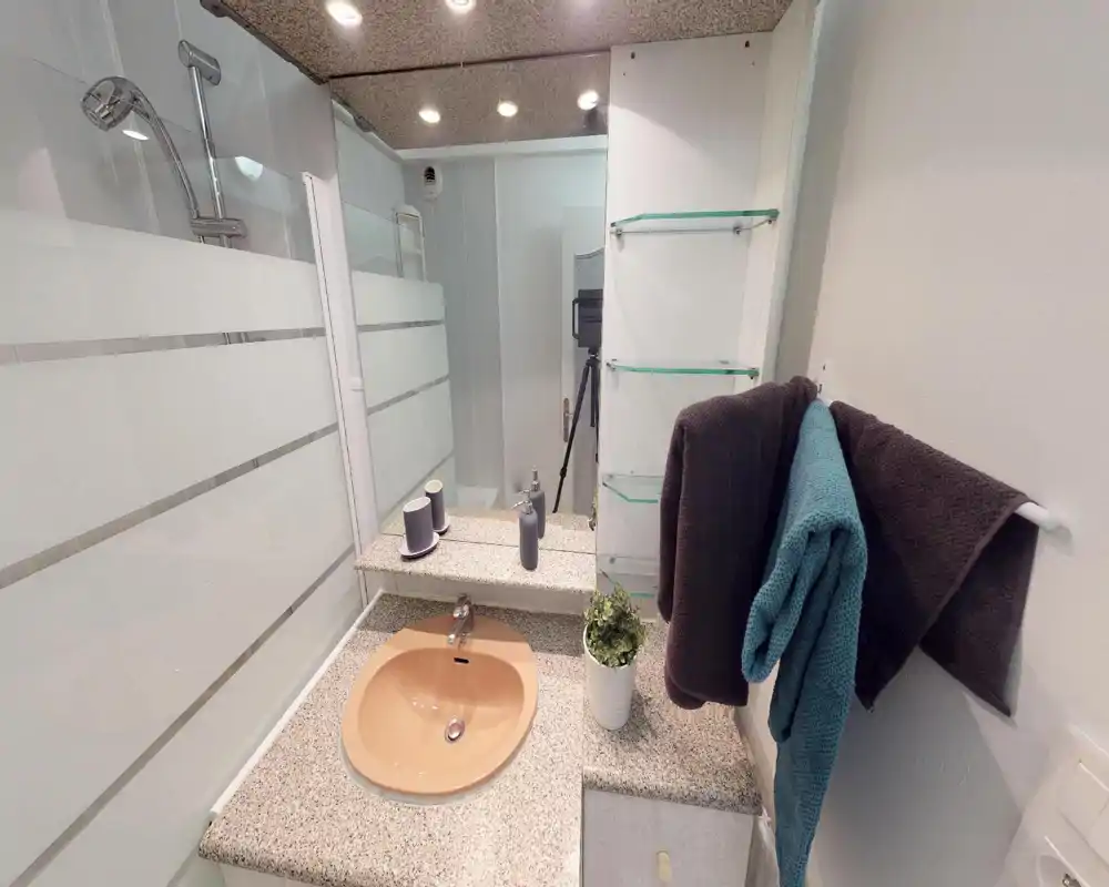 Bagno compatto e ben illuminato con lavabo incassato su piano in granito, specchio, parete doccia in vetro e asciugamani ordinatamente appesi.