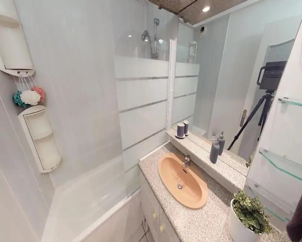 Bagno compatto e moderno con vasca/doccia, lavabo beige incassato in un piano a graniglia, specchio a parete e scaffalature compatte — luminoso e ordinato.