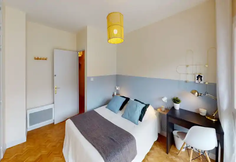 Chambre lumineuse et bien décorée avec un lit double, lampes de chevet et un petit bureau près de la fenêtre. Mur accent bleu et parquet chaleureux créent une ambiance calme et accueillante.